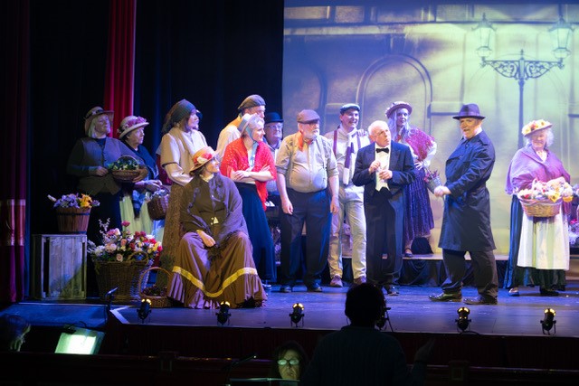 Adrian Bevan (second right) as Higgins asking “Why Can’t the English?” to the Cockneys with Eliza (seated).